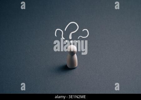 Wooden pawn figurine on dark grey background with hand-drawn question marks above its head symbolizing confusion, uncertainty and decision making. Stock Photo