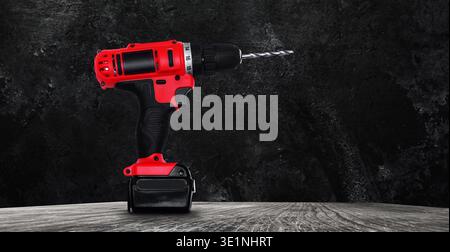 Cordless red power drill with battery standing on a wooden surface against a dark textured wall Stock Photo