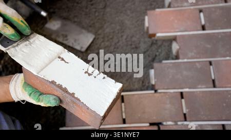 Close up top view of a construction worker applying white mortar to a red brown brick tile with a trowel. Focus on manual labor for paving or home imp Stock Photo