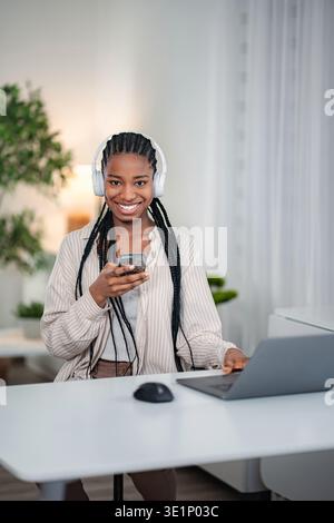 Young woman smiling, using phone and laptop, wearing headphones Stock Photo