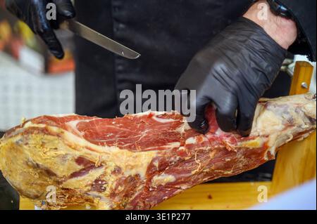 Hand of a professional chef processing a large piece of Jamon on a wooden board. Concept of artisanal meat industry, fresh food supply chain, and skil Stock Photo