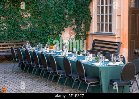 A long table is beautifully set with glassware, plates, and napkins, ready for guests to arrive in a charming, ivy-covered courtyard Stock Photo