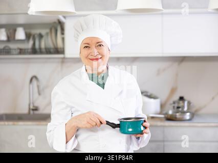 Happy mature female cook in uniform standing and showing her kitchen Stock Photo