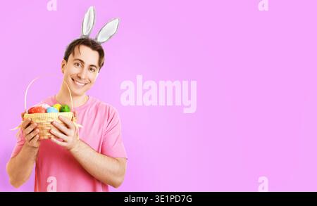 Cheerful man wearing bunny ears holding a basket of colorful Easter eggs and smiling at camera against a bright pink background with ample copy space Stock Photo