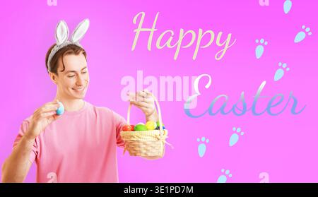 Cheerful man in bunny ears holding a blue Easter egg and a basket full of colorful eggs against a pink background with Happy Easter text and paw print Stock Photo