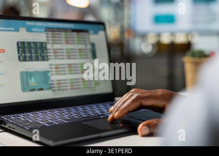 Financial analyst at desk evaluating investment opportunities and company stock performance on laptop. African American office employee studying online market projections and data on device. Stock Photo