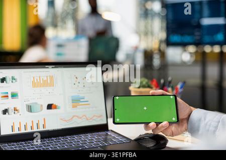 Male professional grasping mobile phone with green screen beside laptop on office desk, checking financial charts. Business analyst working in modern office using isolated chroma key display. Stock Photo