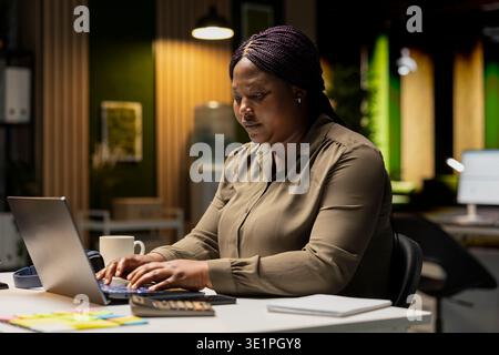 Female specialist working after hours on proofreading and fact checking, writing important data on her laptop. Creating a summary and designing strategy to align with company targets. Stock Photo