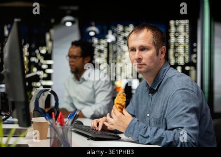 Developer eating croissant in office, reviewing system metrics, monitoring performance dashboards. Tech company IT expert enjoying snack at desk, tuning neural networks using program on computer Stock Photo