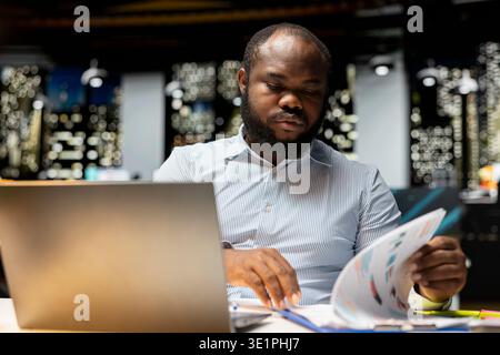 Black advisor researching numbers and finance figures in a dark office at midnight, documenting updates and organizing the agenda for upcoming business briefing. Setting targets at night. Stock Photo