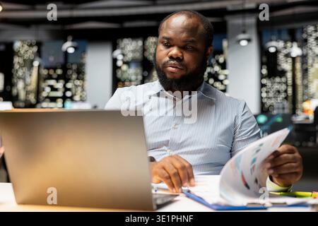African american businessman verifying numbers and finance data in a dark office after hours, documenting updates and fixing the targets for upcoming business activity. Proofreading at night. Stock Photo