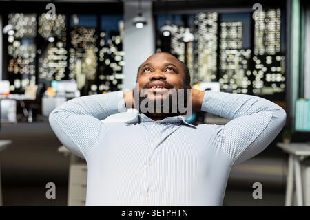 Fulfilled black businessman enjoys a quiet moment of downtime in a modern office, reclining in his desk chair with a comfortable and laid back posture. Resting after tasks, empowerment. Stock Photo