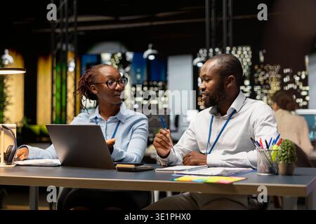 Manager helping young intern in office to complete spreadsheets using laptop, analyzing data. Team leader assisting trainee researching information online on notebook, working overnight Stock Photo