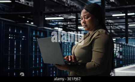 Software developer in data center using laptop to upgrade systems for increased stability. Server hub employee configuring equipment rigs with notebook for optimum performance, camera A Stock Photo