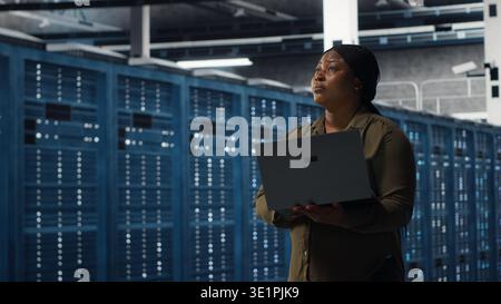 Smiling engineer walking in data center with laptop, checking hardware for system stability. Upbeat employee overseeing server farm with notebook, configuring infrastructure, camera B Stock Photo