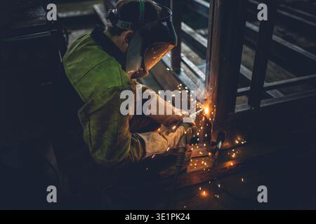 A medium side shot of a welder in a protective mask and heavy-duty suit performing metal arc welding. Sparks are flying from the contact point. The da Stock Photo