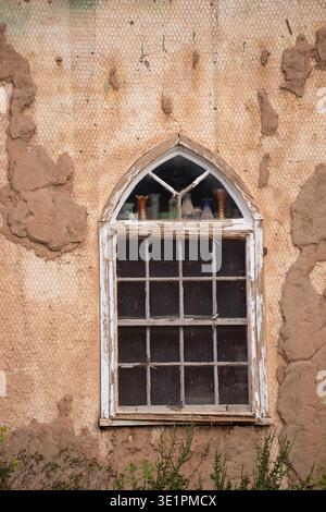 antique arch shaped window in old building under repair wall with plaster still clinging to the mesh window has antique bottles on the window ledge Stock Photo