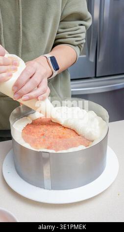 Individual in a green sweatshirt decorates a layered cake with cream frosting from a piping bag, positioned on a white cake stand in a contemporary ki Stock Photo