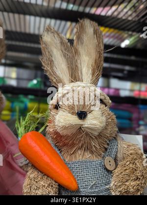 Straw Bunny Holding Bright Carrot, Knitted Sweater With Button Detail, Rustic Market Display On Shelf Offering Playful Farmhouse Easter Vibe With Hand Stock Photo