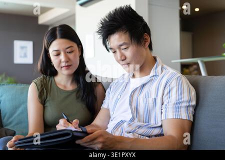 Asian couple sitting on gray sofa in living room reviewing black portfolio and signing with pen Stock Photo