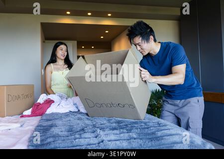 Asian couple packing donations on bed at home with cardboard boxes and folded clothes Stock Photo
