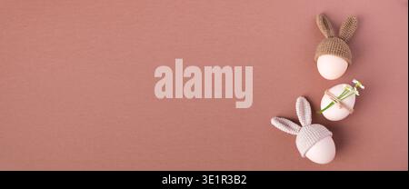 Three decorative Easter eggs wearing knitted bunny hats, one adorned with a flower, arranged on a soft pink background for festive decoration Stock Photo