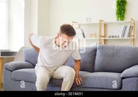 Sick man with lower back pain sitting on sofa at home, suffering from backache Stock Photo