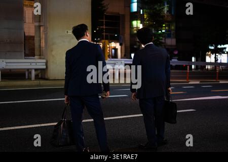 Two Japanese salarymen in business suits standing on a street corner at night in Roppongi, Tokyo, holding briefcases while waiting to hail a taxi. Stock Photo