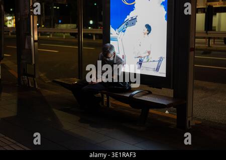 An elderly man wearing a face mask sleeps on a bench at a bus stop at night in Tokyo, illuminated by the bright glow of a backlit advertising display. Stock Photo