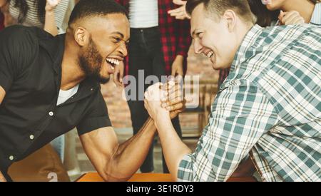 Two Men Engage in an Arm Wrestling Match Surrounded by Friends in a Social Gathering Setting Stock Photo