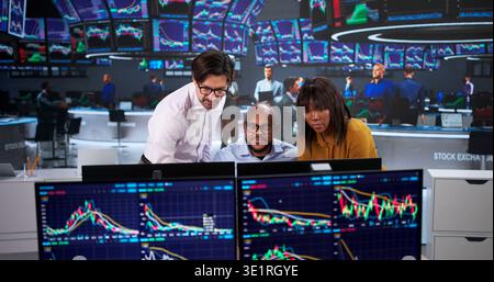 Diverse Group Of Financial Brokers And Agents Monitoring Stock Market Charts On Computers, Discussing Reports Stock Photo