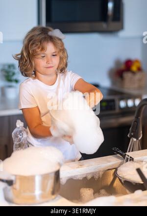 Boy washing dishes with bubbles. Child playing with foam in kitchen. Cute kid cleaning dishes at home. Happy child splashing soap foam. Funny boy Stock Photo