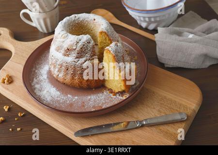 Homemade bundt cake with powdered sugar on wooden cutting board. Cozy kitchen scene featuring ceramic baking mold, wooden spoon, linen napkin and cup Stock Photo
