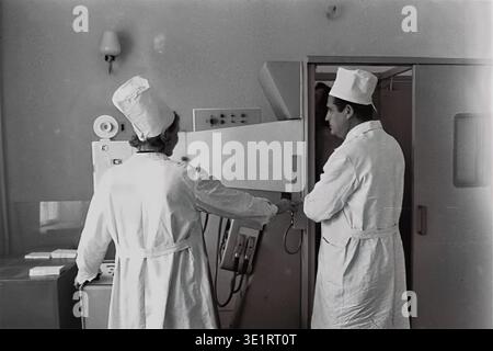 A black and white archival photograph taken in 1977 depicts two medical workers operating a large diagnostic X-ray or fluorography machine at the AIZ factory hospital in the city of Slavyansk. A male and a female doctor in white coats and tall medical caps stand by the control panel, while a patient is positioned inside the imaging booth. This scene highlights the diagnostic technology of Soviet healthcare. This historical picture documents preventive occupational medicine and peaceful Donbas before the war. Stock Photo