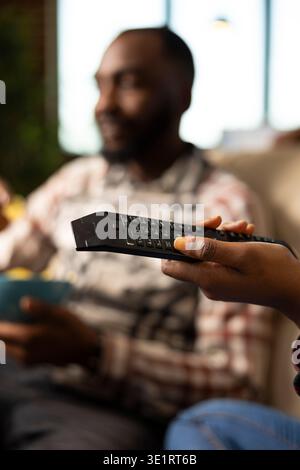 African american couple on couch, bonding over movies at home. Closeup shows black woman using remote to adjust volume or change channel while man relaxes with snacks in background. Stock Photo