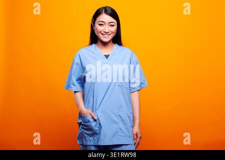 Young woman in medical scrubs, having hand in a pocket and posing in front of yellow background. Portrait of female asian nurse standing and smiling at camera, portraying readiness for healthcare. Stock Photo