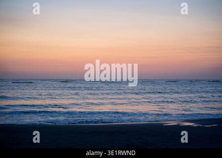 Soft pastel orange and pink twilight sky over calm ocean waves in Tainan, Taiwan, with distant fishing net silhouettes lining the serene horizon. Stock Photo