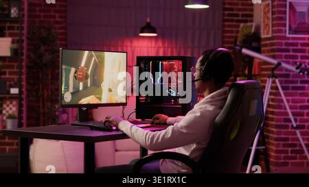 Static tripod shot of african american gamer streaming first person shooter while explaining gameplay while using pc gaming setup. Man relaxing playing online action game talking to team on headset. Stock Photo