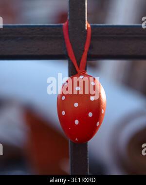 Red egg decorated with white dots is suspended from a black metal crossbar, with a softly blurred background of colorful decorative objects Stock Photo