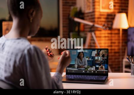 African American female manager leading virtual team discussion on laptop from cozy home during sunset. Black businesswoman attends video conference call, portraying remote work productivity. Stock Photo
