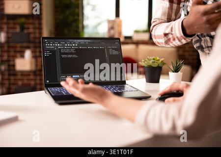 Closeup on laptop displaying programming language as multiethnic people discuss on building an app from home. Diverse couple brainstorming and debugging code for web development in modern apartment. Stock Photo