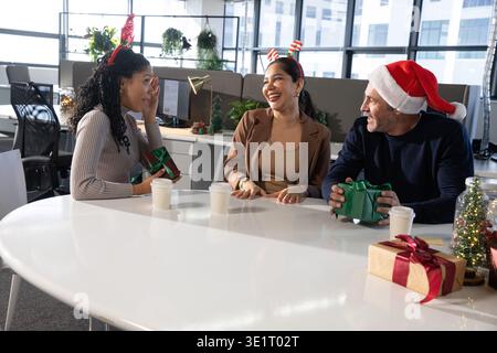 Diverse coworkers exchanging gifts sitting at round white table in office, wearing festive headwear Stock Photo