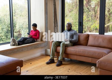 African American father and boy sitting at home on tan sofa using silver laptop and smartphone Stock Photo