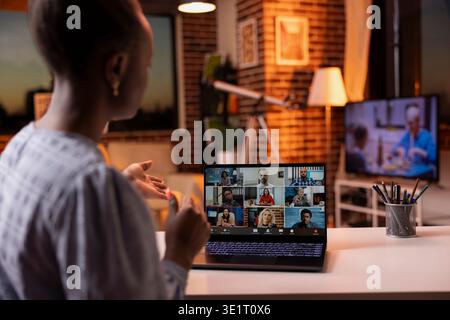 Young black woman working remotely from cozy living room during sunset, attending online video conference. Female entrepreneur having virtual discussion with diverse coworkers on laptop. Stock Photo