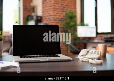 Empty startup office with open laptop, telephone, papers and coffee cup neatly placed on desk. Modern brick wall workspace having digital device with black screen on vacant table. Stock Photo