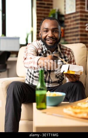 Casual moment of black male enjoying fast food and watching TV from couch. Happy man seated in modern apartment, pointing remote at screen while looking for show or movie to watch. Stock Photo