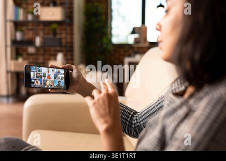 Woman entrepreneur seated with phone in hand, discussing with multiethnic professionals in online workshop. Close up of female manager using smartphone for video call with remote colleagues. Stock Photo