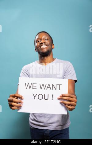 African American male recruiter holding a hiring ad, searching for potential candidates to support company expansion. Smiling black businessman in casual attire promotes workforce development. Stock Photo