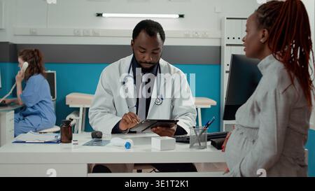 General practitioner taking notes on digital tablet at maternity checkup with pregnant patient. Male physician having conversation about pregnancy with future mother in medical office. Stock Photo