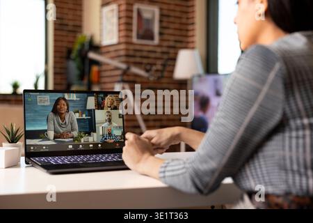 Focused female manager engages in online discussion from home, addressing workplace concerns through laptop video conference with distant coworkers. Virtual meeting among diverse professionals. Stock Photo